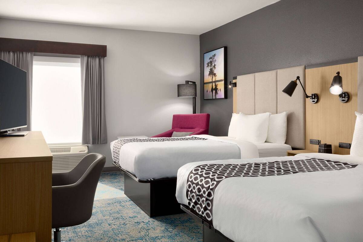 Une chambre d’hôtel avec deux lits, un bureau, une chaise, une télévision et une grande fenêtre avec des rideaux.