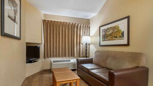 A hotel room with a brown leather sofa, a wooden table, a TV, a lamp, and a window with curtains. - Extended Stay America Suites Indianapolis West 86th St (Indianapolis)