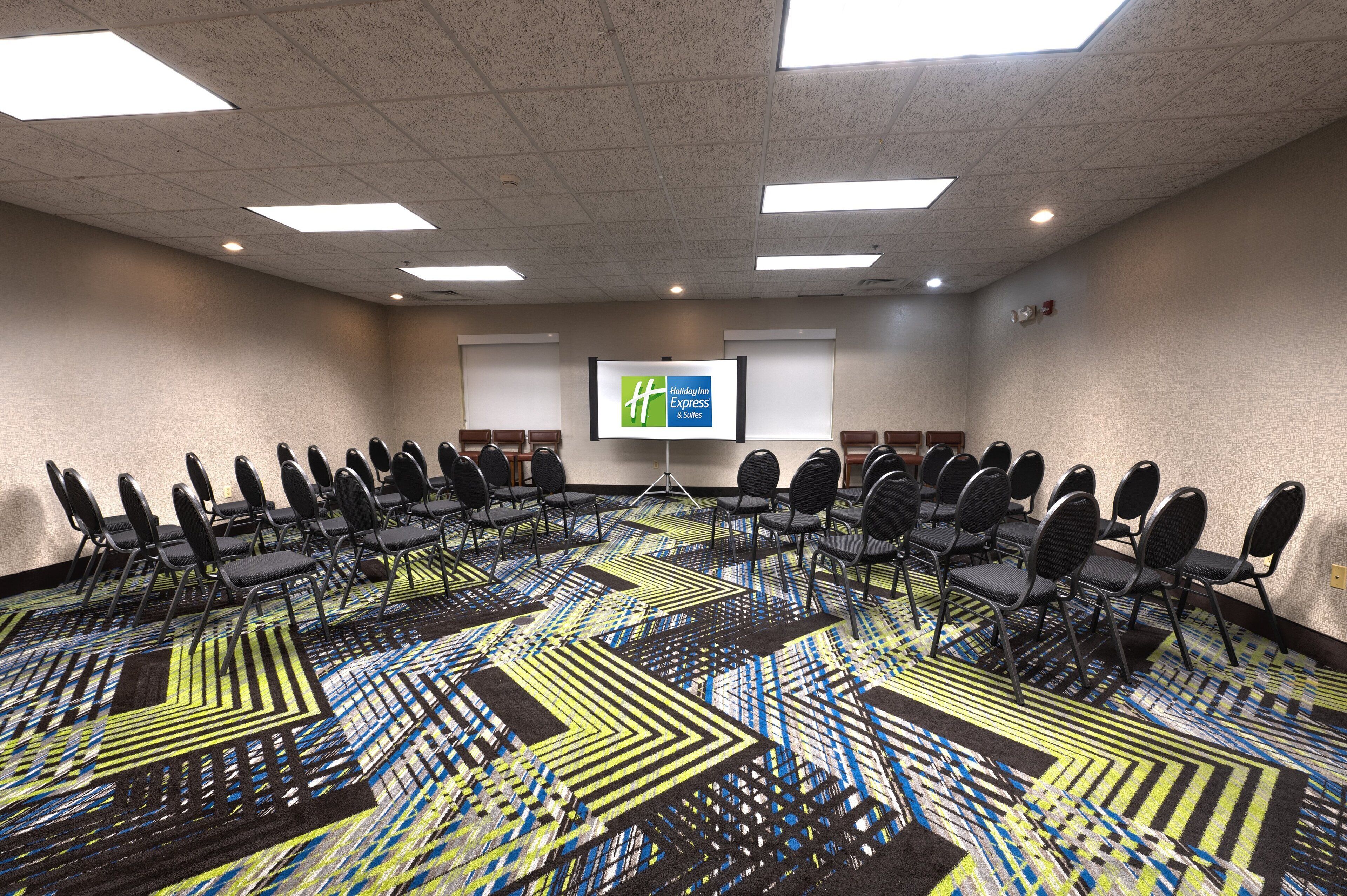 A conference room with a patterned carpet, rows of chairs, a projector screen, and a presentation screen.
