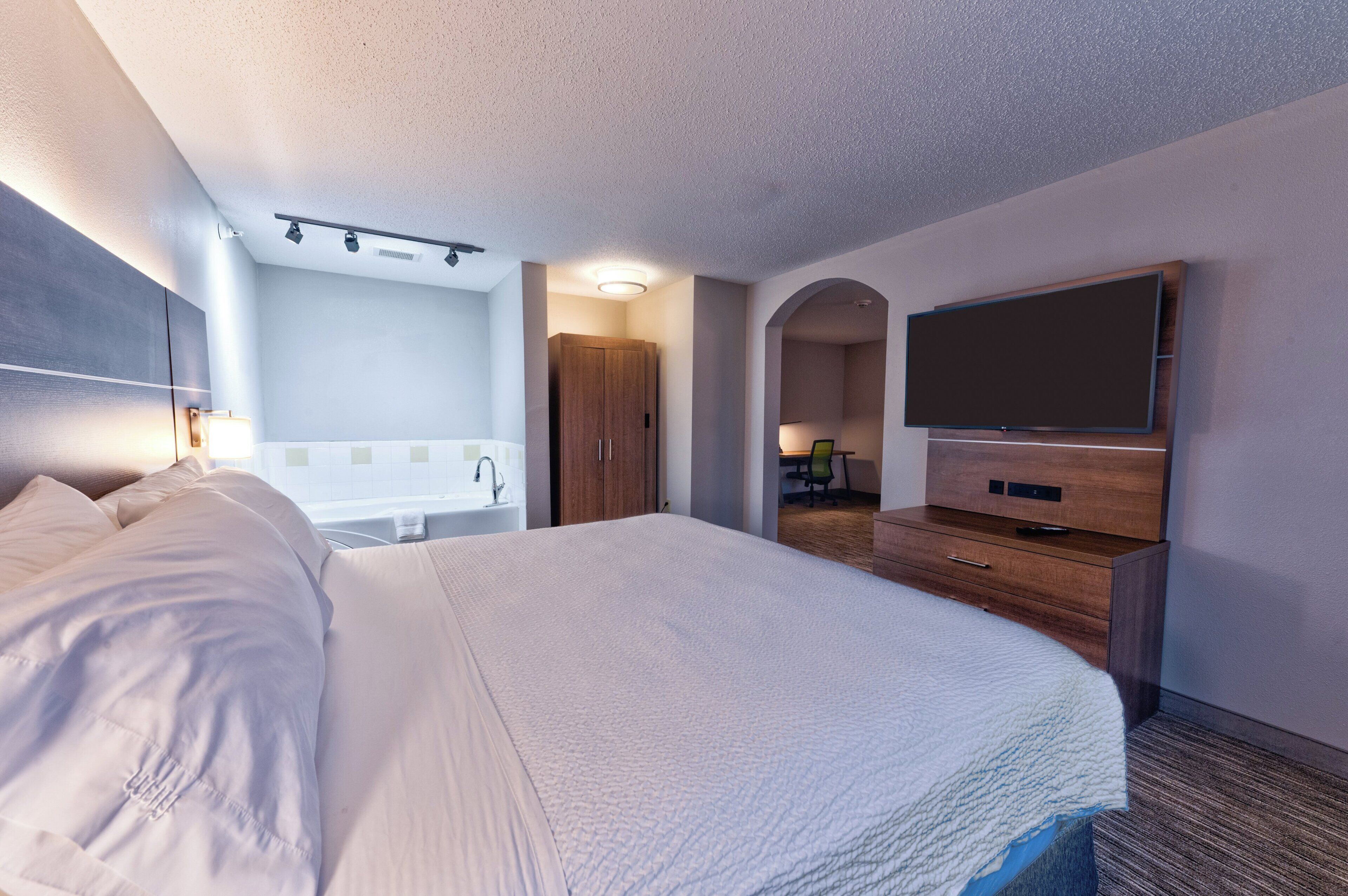 A hotel room with a large bed, a flat-screen TV, a wooden dresser, a bathroom with a bathtub, and a desk with a chair.