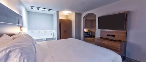 A hotel room with a large bed, a flat-screen TV, a wooden dresser, a bathroom with a bathtub, and a desk with a chair.