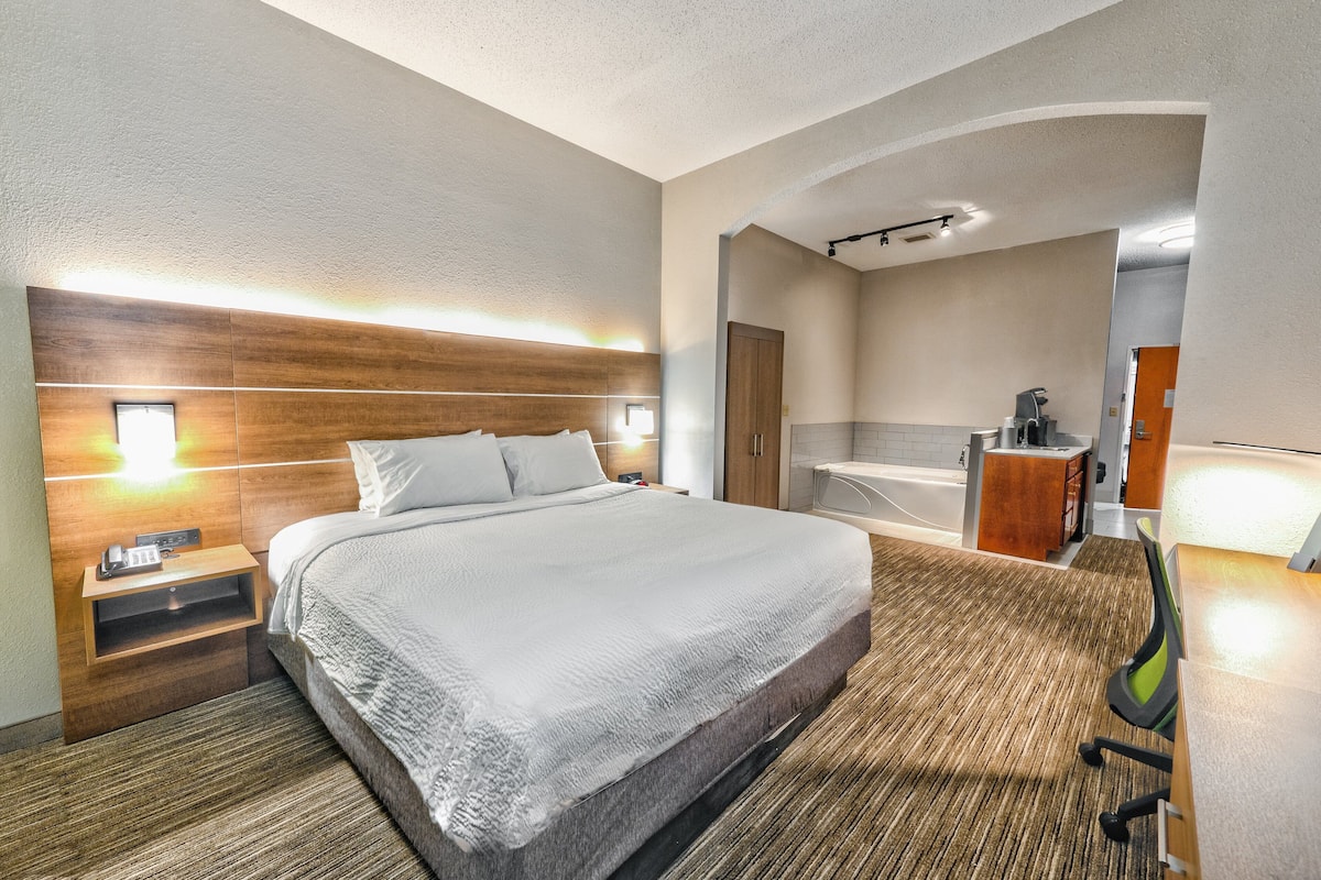 A hotel room with a large bed, bedside table with a phone, a nightstand, a bathroom with a bathtub, and a shower area.