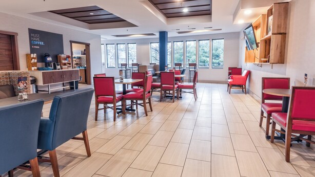 Un área de comedor moderna con sillas rojas y azules, una estación de café y amplias ventanas que ofrecen luz natural.