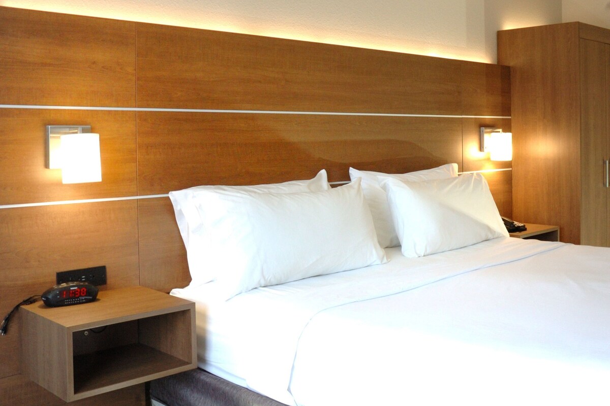A hotel room with a wooden headboard, two bedside lamps, a nightstand with a clock, and a large bed with white bedding.