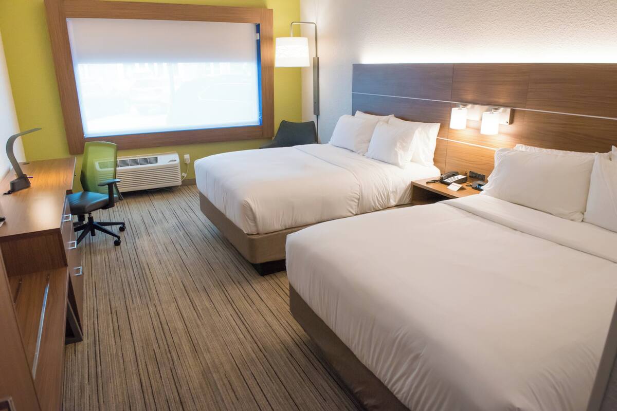 A hotel room with two beds, a desk, a chair, a lamp, and a window with blinds.