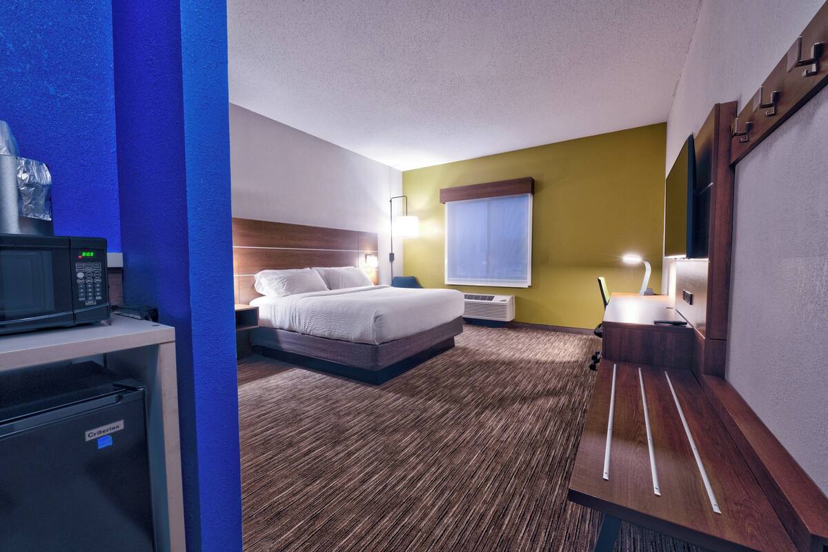 A hotel room with a bed, bedside table, lamp, and a microwave on a counter.