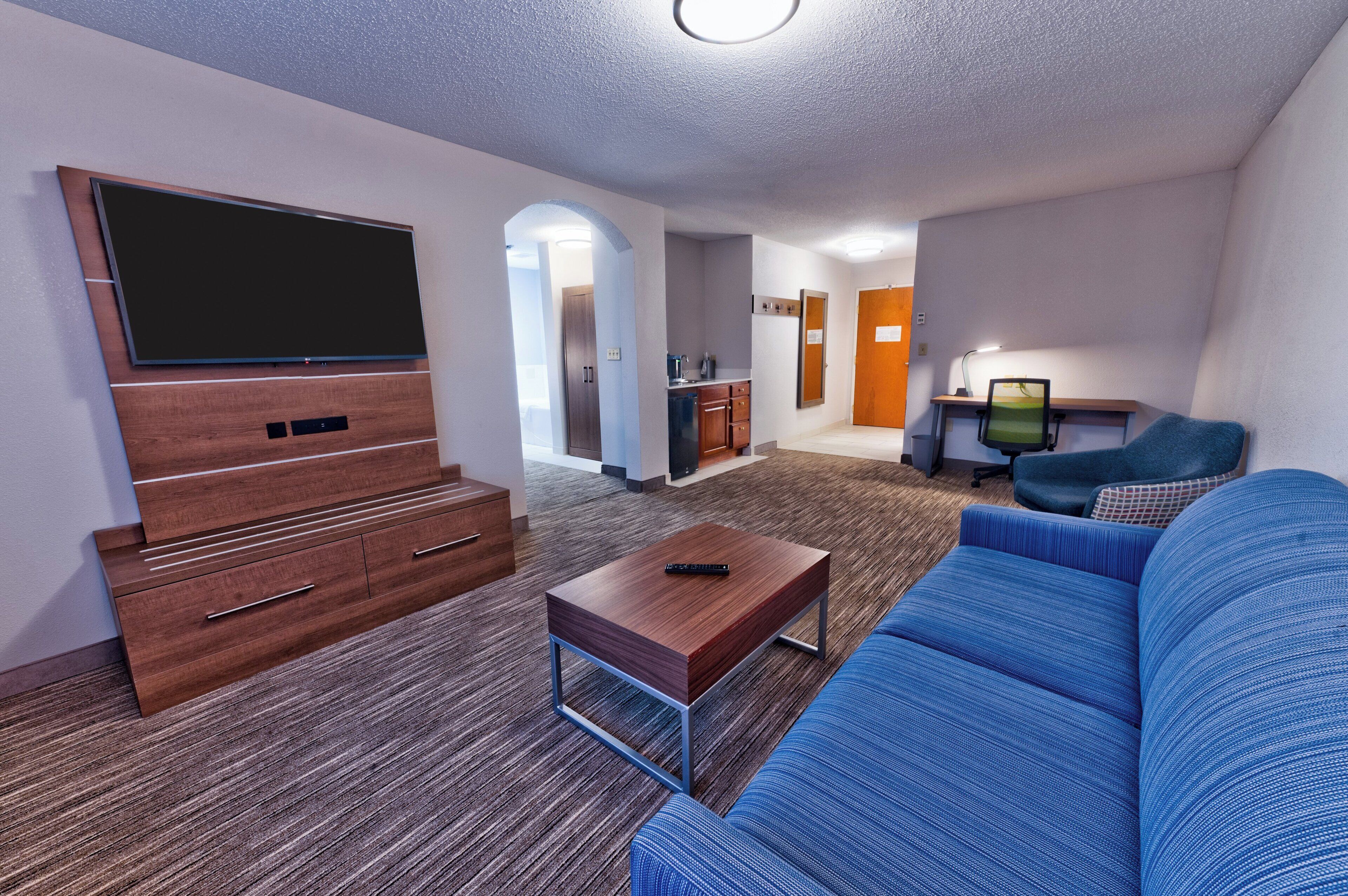 A modern living room with a flat-screen TV, a blue sofa, a wooden coffee table, and a kitchen area in the background.
