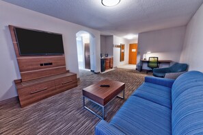 A modern living room with a flat-screen TV, a blue sofa, a wooden coffee table, and a kitchen area in the background. - Holiday Inn Express Hotel & Suites Goshen by IHG (Goshen)