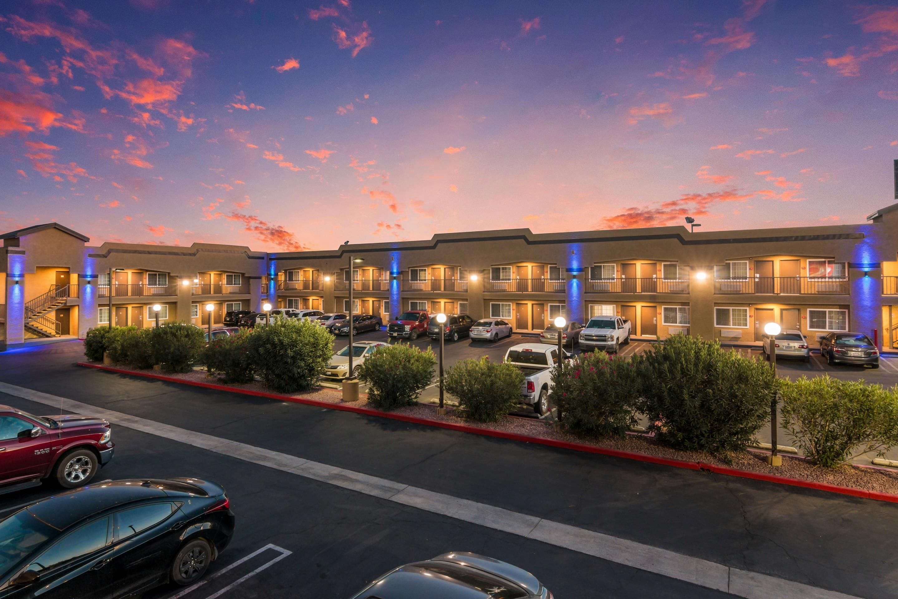 A multi-story motel with a parking lot, featuring a row of rooms with balconies and a clear sky.