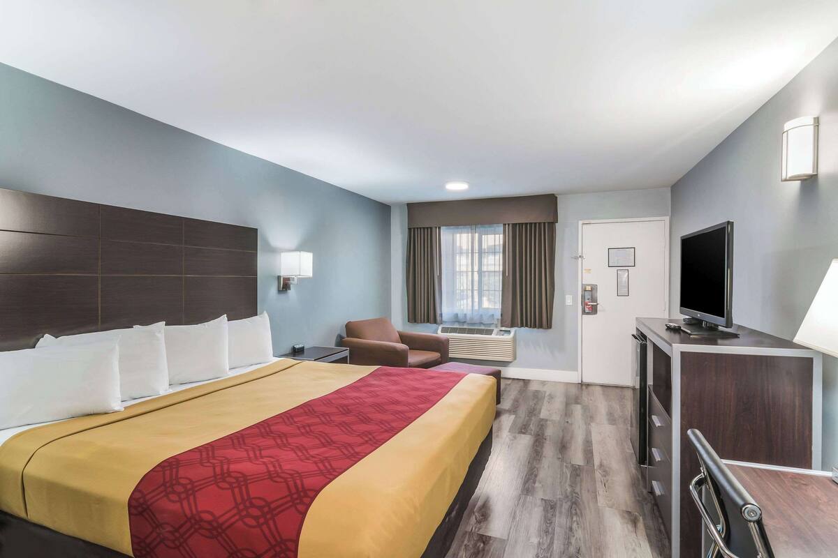 A hotel room with a large bed, a desk with a flat-screen TV, a chair, and a window with curtains.