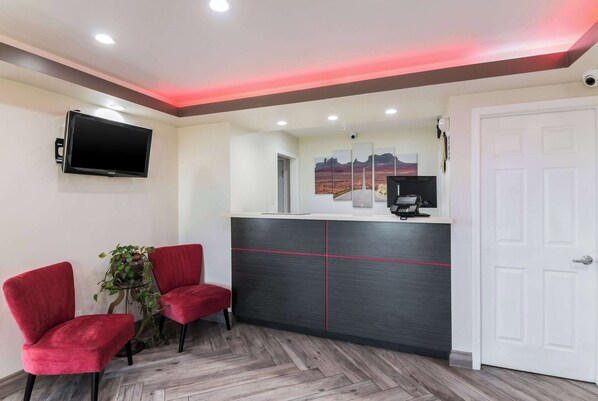 A reception area with a reception desk, two red chairs, a potted plant, a mounted TV, and a door. - Econo Lodge Hesperia - Victorville I-15 (Hesperia)