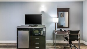 Habitación de hotel con escritorio, una silla, televisión de pantalla plana, microondas, refrigerador, lámpara y espejo.