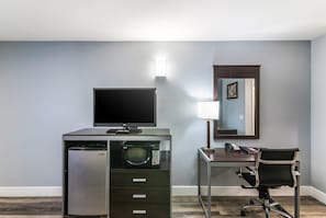 A hotel room with a desk, a chair, a flat-screen TV, a microwave, a refrigerator, a lamp, and a mirror.
