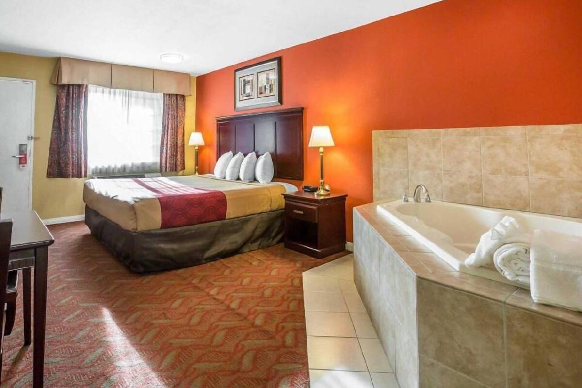 A hotel room with a bed, bedside table, lamp, and a large bathtub.
