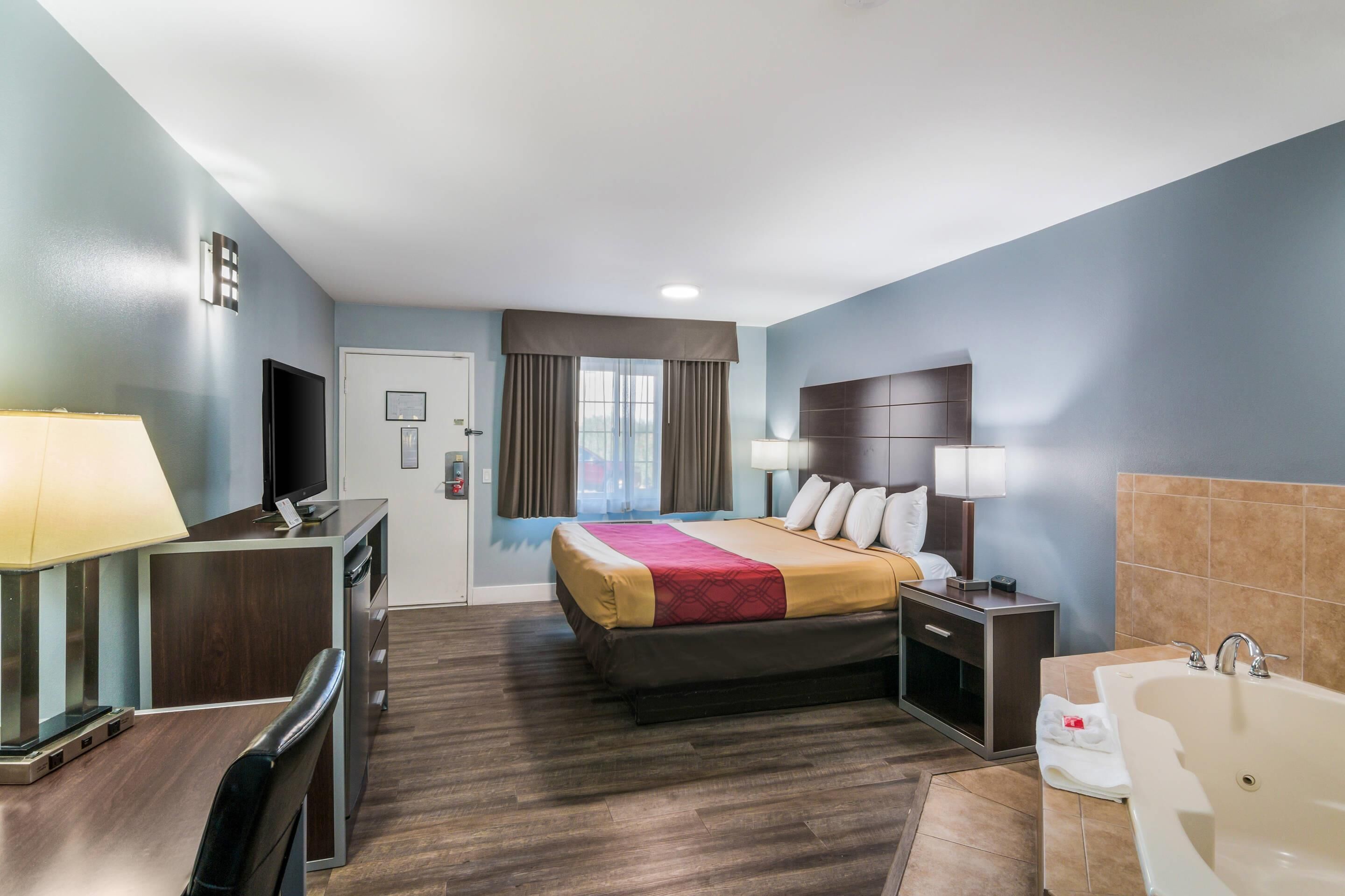 A hotel room with a bed, a desk with a computer, a television, a bathroom with a bathtub, and a window with curtains.