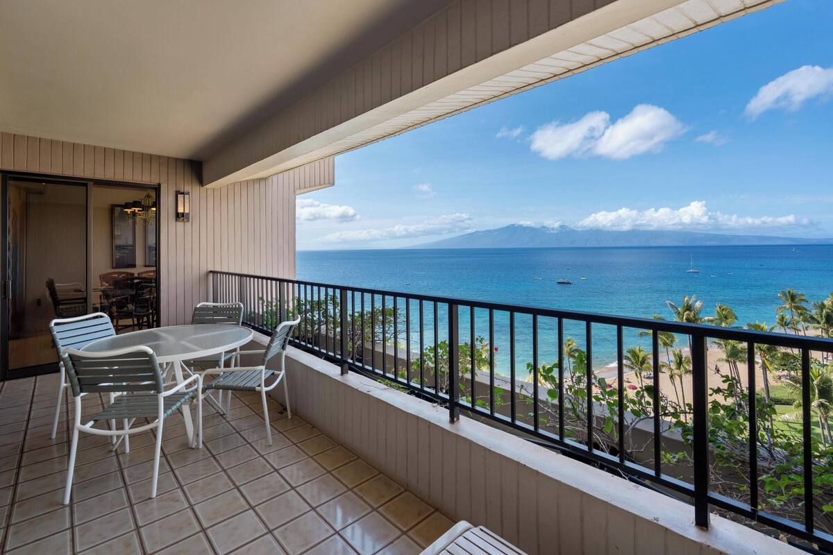 A balcony with a table and chairs, a view of the ocean, and a distant mountain range.