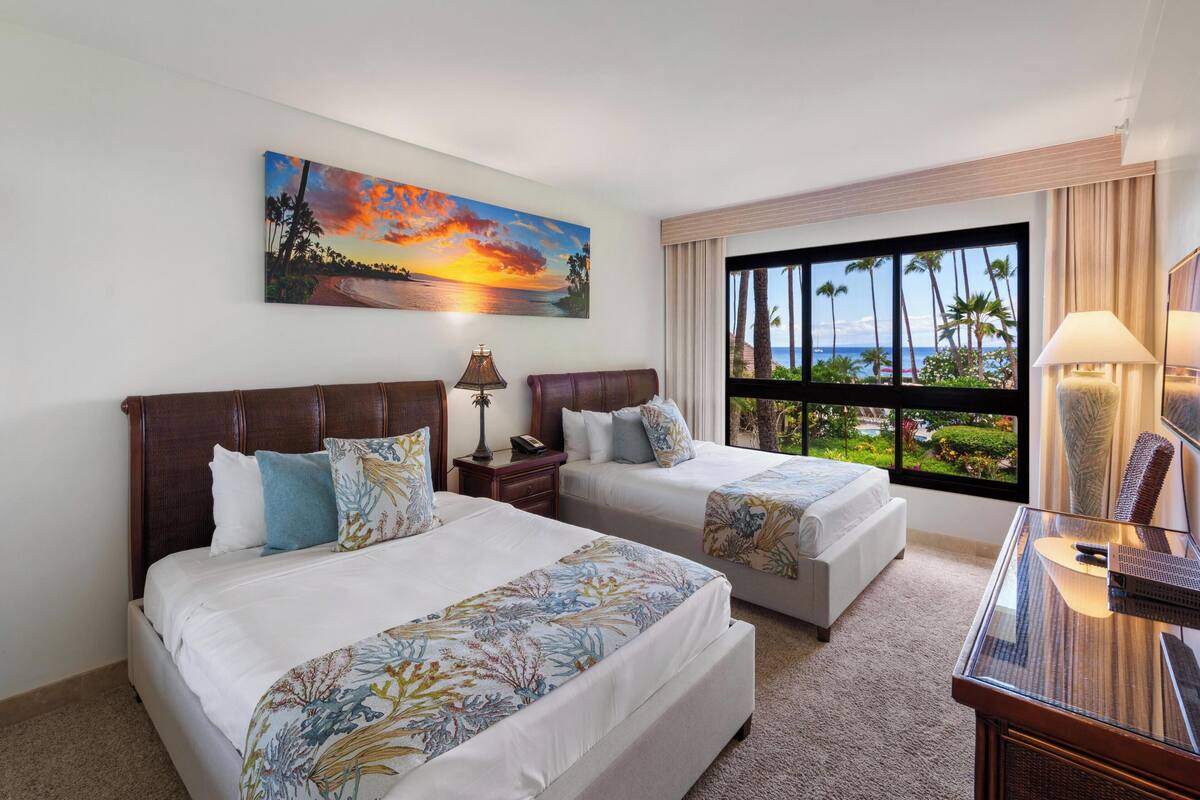 A hotel room with two beds, a painting of a sunset, a large window with a view of palm trees and a beach, and a desk with a lamp.