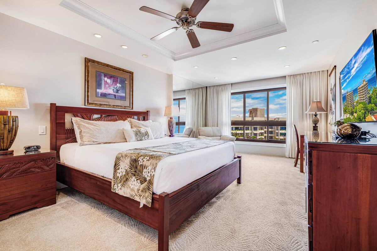 A hotel bedroom with a large bed, a ceiling fan, a TV, and a view of the cityscape through the window.