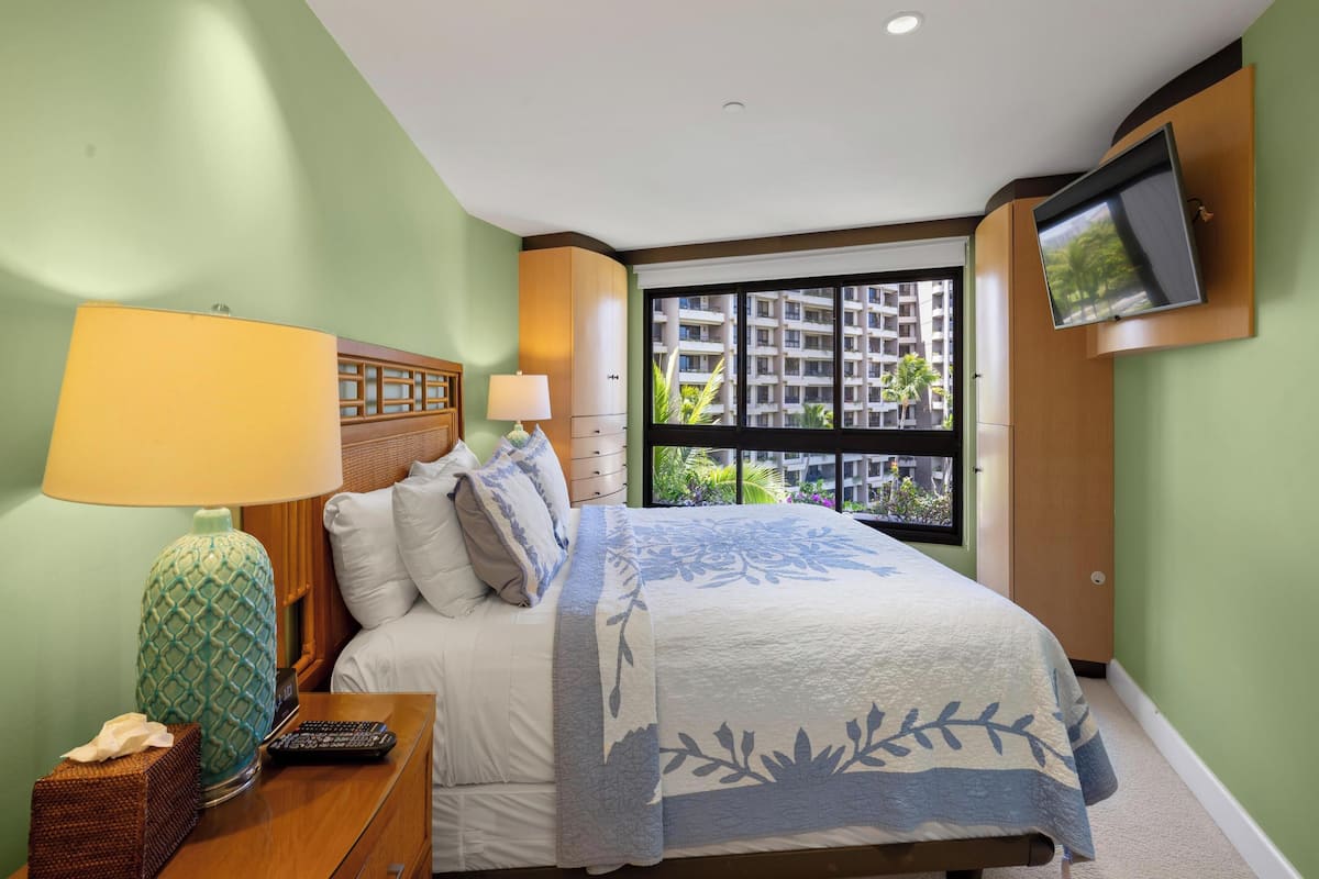 A hotel room with a bed, a TV mounted on the wall, a window with a view of buildings, and a nightstand with a lamp and remote control.