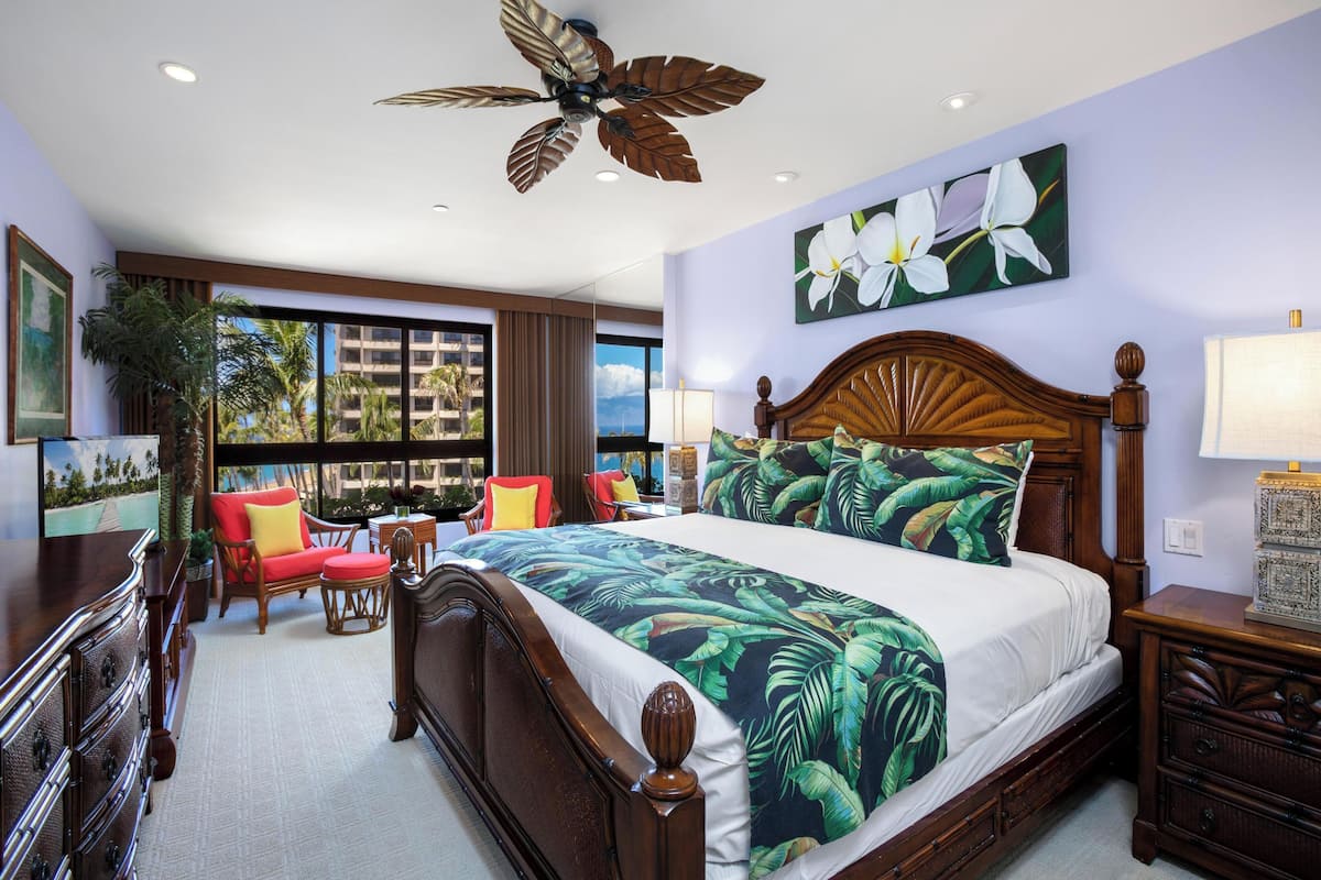 A hotel room with a large bed, a wooden headboard, a ceiling fan, a window with a view of palm trees, and a red chair.