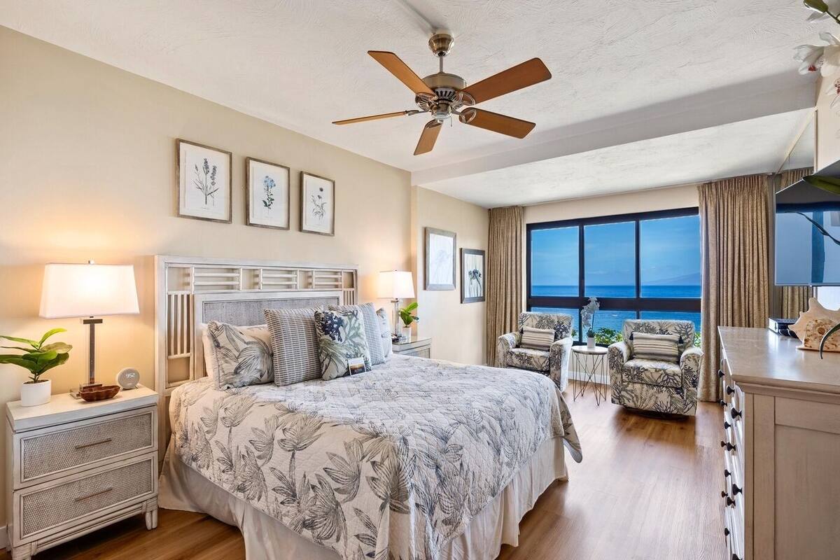 A bedroom with a bed, nightstands, a ceiling fan, framed botanical prints, and a view of the ocean.