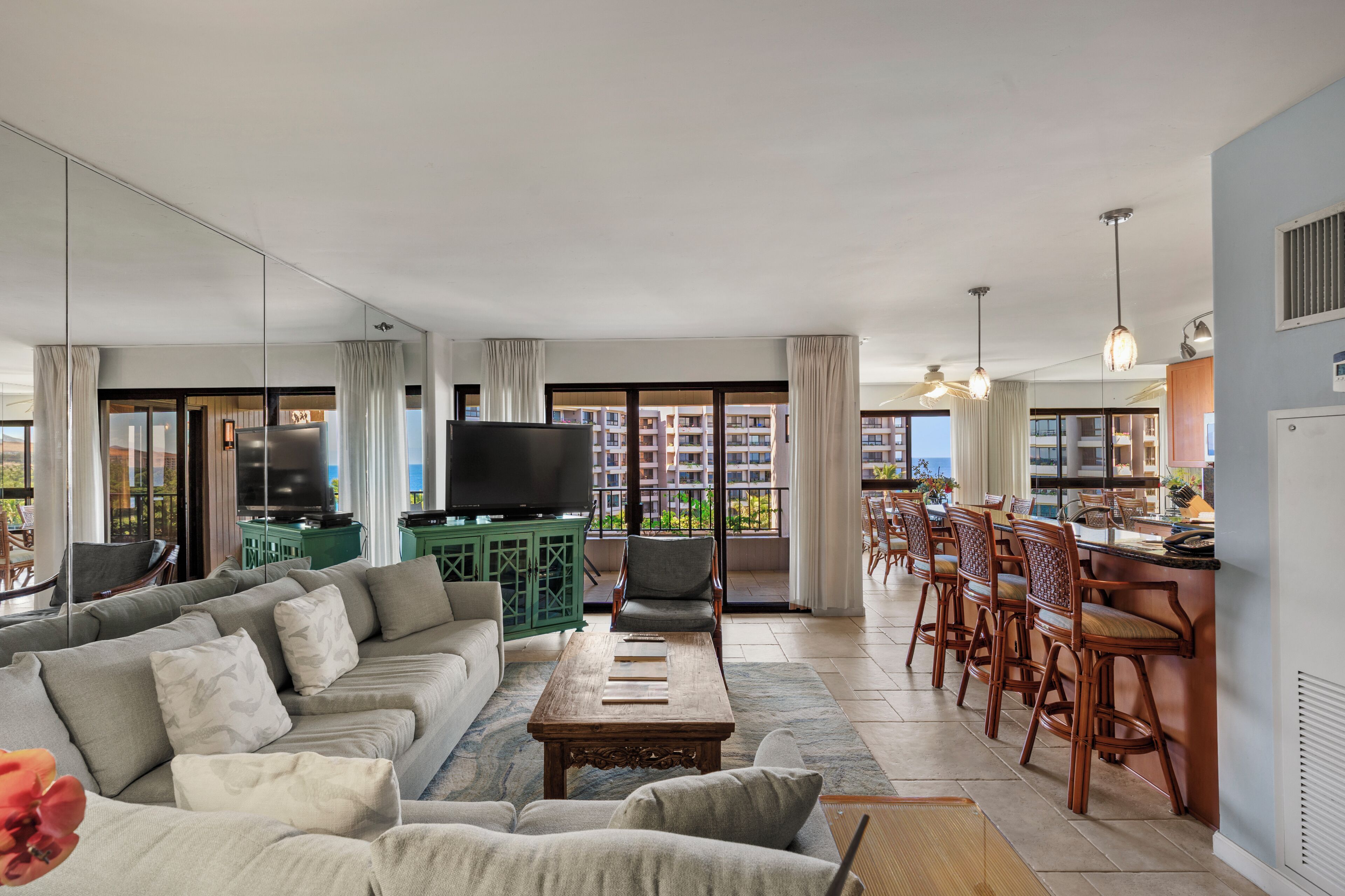 A spacious living room with a large sofa, a coffee table, and a TV. There is a balcony with a dining area and a kitchen.