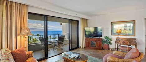 Un vaste salon doté d’une grande porte-fenêtre coulissante donnant sur un balcon avec vue sur l’océan. On y trouve un téléviseur, un fauteuil en osier et un canapé avec des coussins.