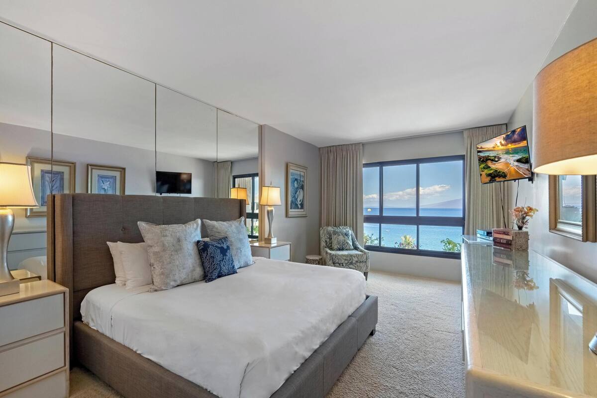 A hotel room with a large bed, a desk, a chair, a TV, and a view of the sea.