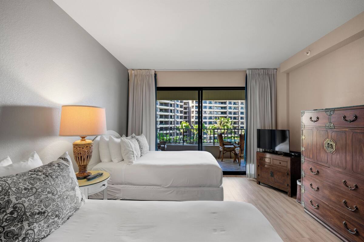 A hotel room with a bed, a TV, a dresser, and a view of a building through the window.