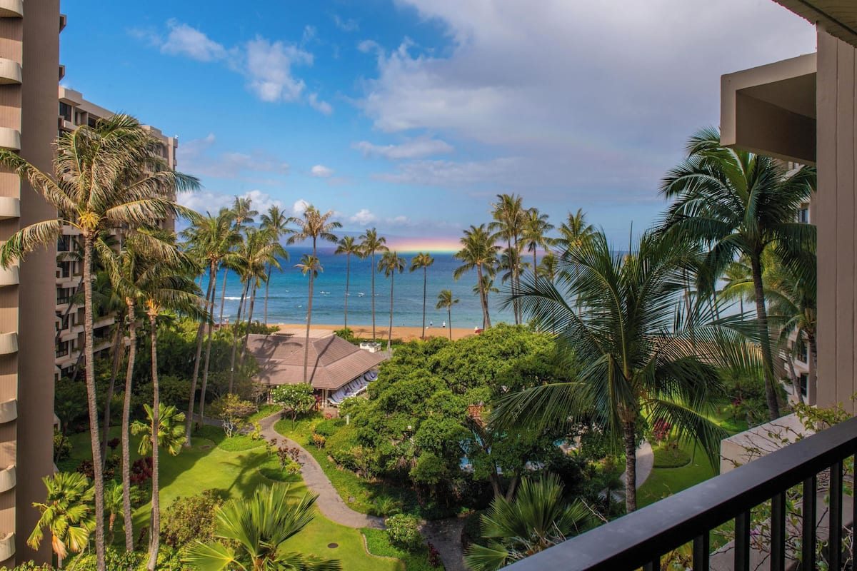 A hotel balcony with a view of a beach, palm trees, and a clear sky.