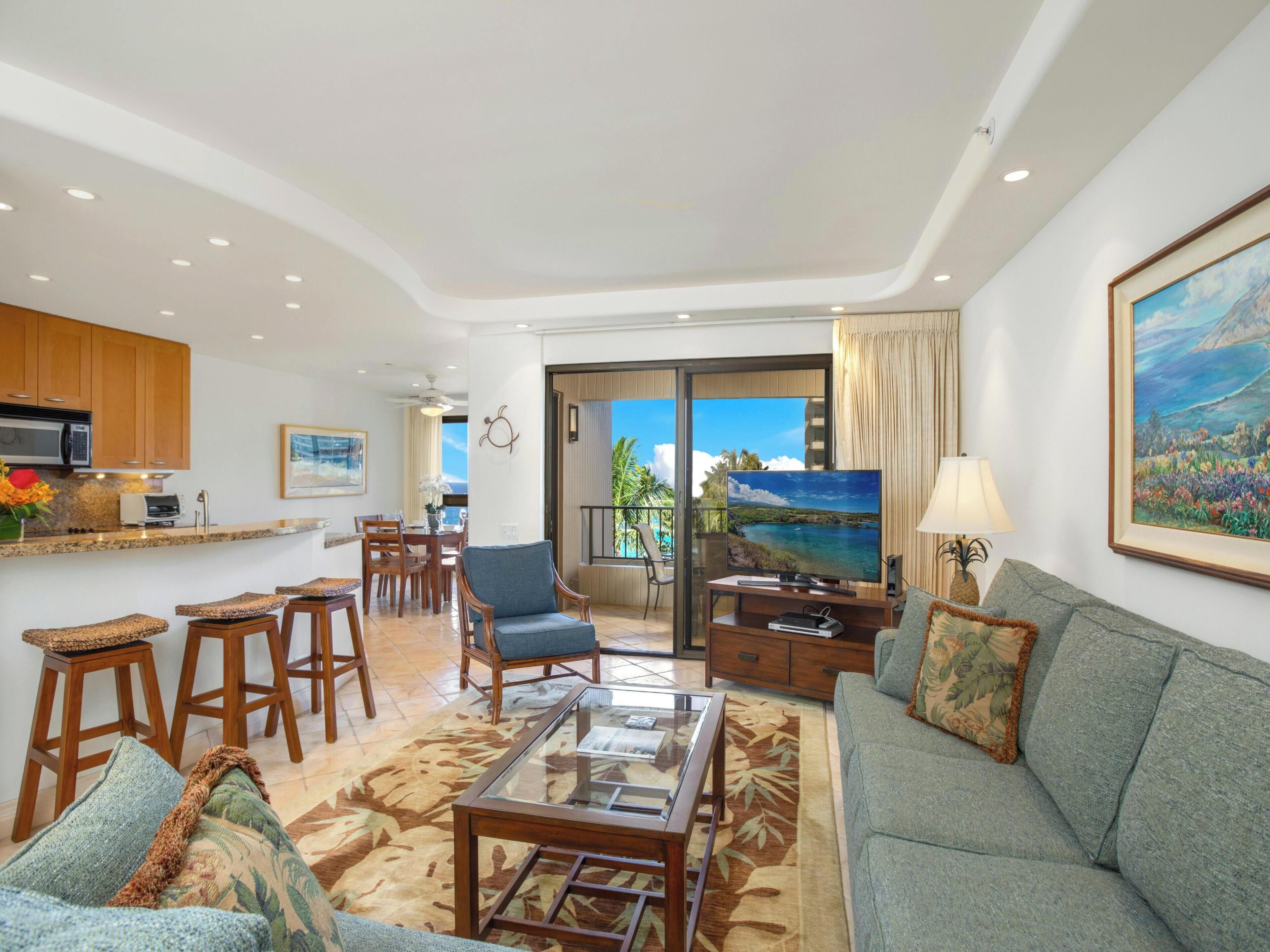 A spacious living area with a sofa, a coffee table, and a TV. There is a balcony with a view of the ocean.