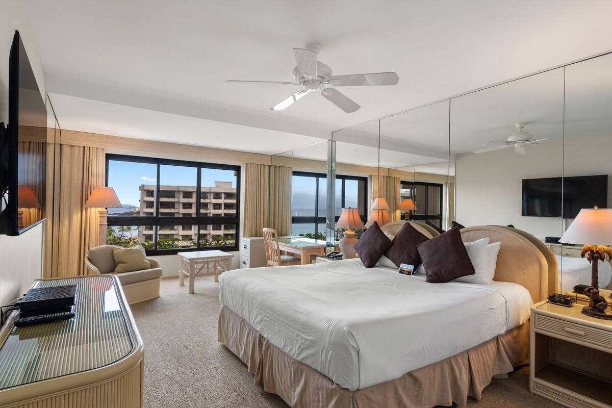 A hotel room with a large bed, a desk, a chair, a TV, and a ceiling fan.