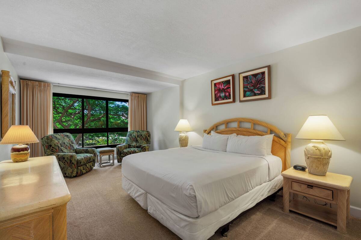 A hotel room with a large bed, two bedside lamps, a nightstand, a chair, a small table, and a window with curtains.