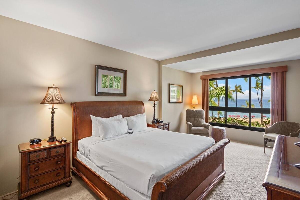 A hotel room with a large bed, two bedside tables, a chair, a desk, and a window with a view of the beach.