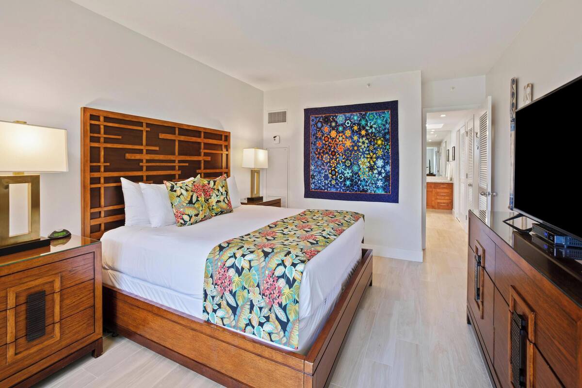 A hotel room with a wooden headboard, a large bed with floral bedding, a nightstand with a lamp, a TV, and a wall art piece.