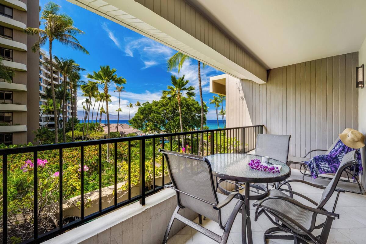 A balcony with a table and chairs, overlooking a beach with palm trees and a hotel building.