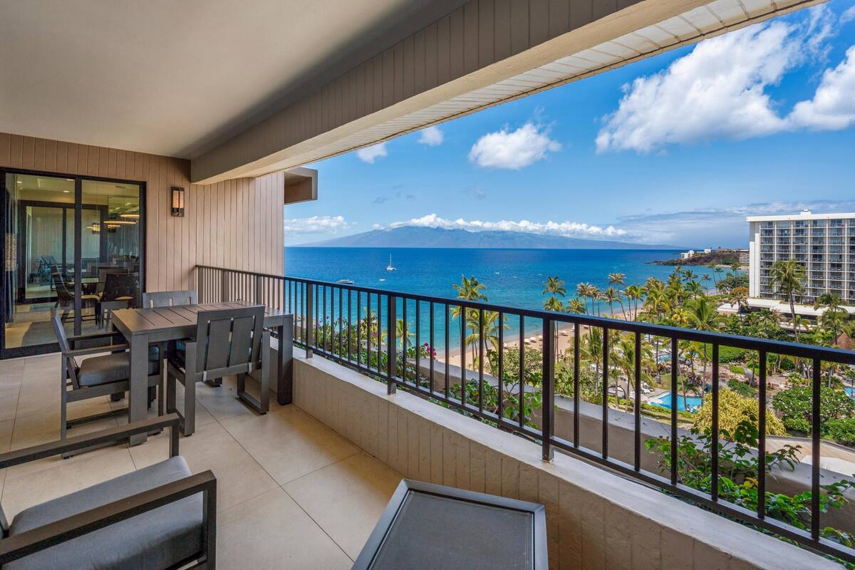 A balcony with a view of the ocean, a dining area with tables and chairs, and a building in the background.