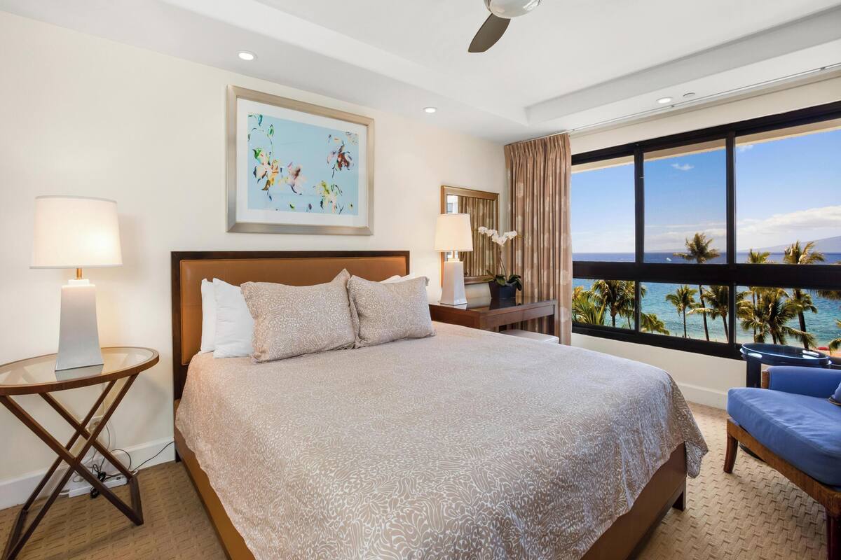 A hotel room with a large bed, a ceiling fan, a view of the ocean, and a painting on the wall.