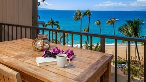 A wooden table with a book, a white mug, and a glass jar with a rope, set against a balcony overlooking a beach with palm trees and the ocean. - Kaanapali Alii-2 Bedroom Ocean Views (Lahaina)