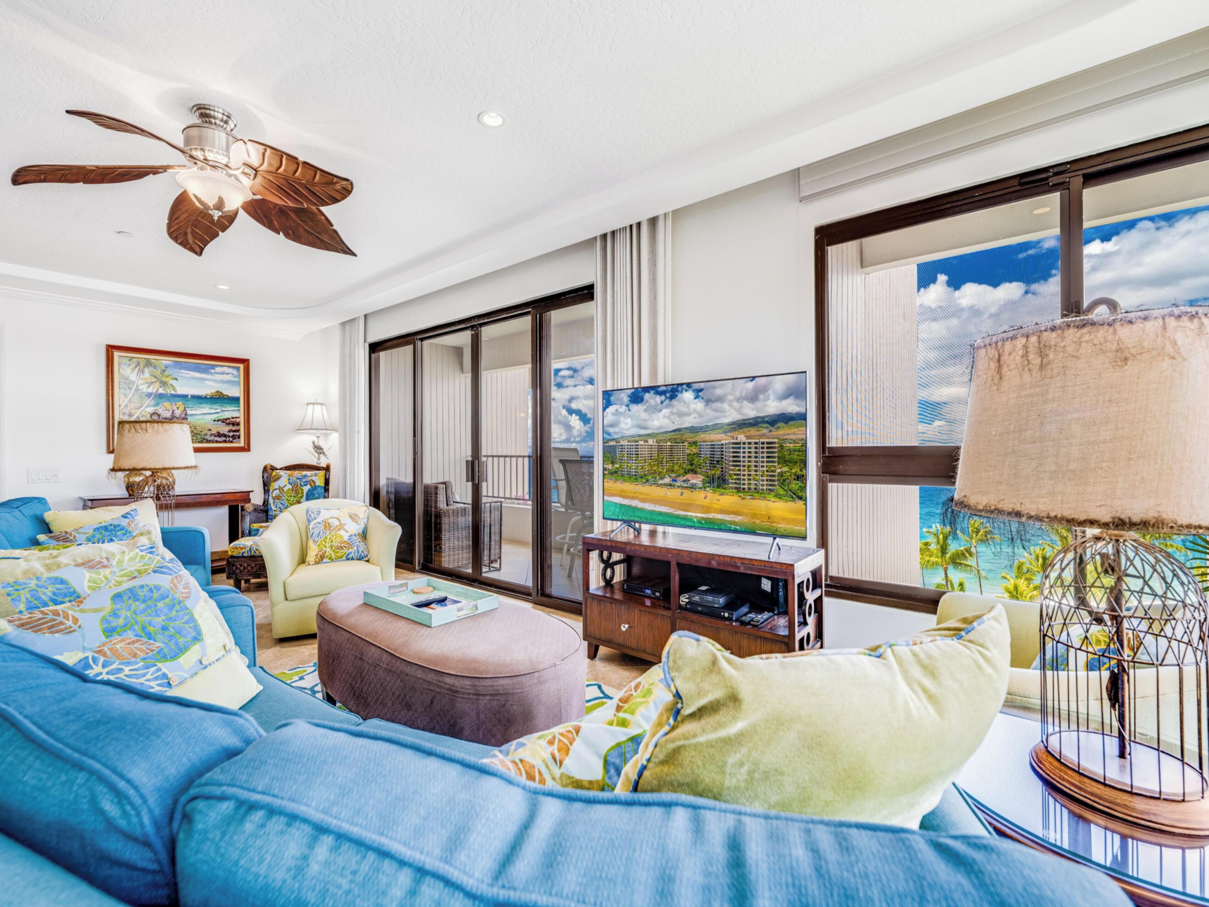 A spacious living room with a large flat-screen TV, a ceiling fan, and a view of the beach through large windows.