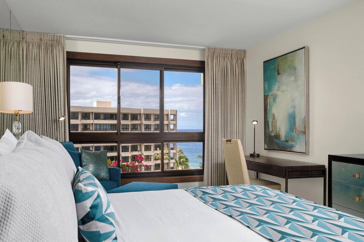 A hotel room with a large bed, a desk, and a view of a building and ocean.