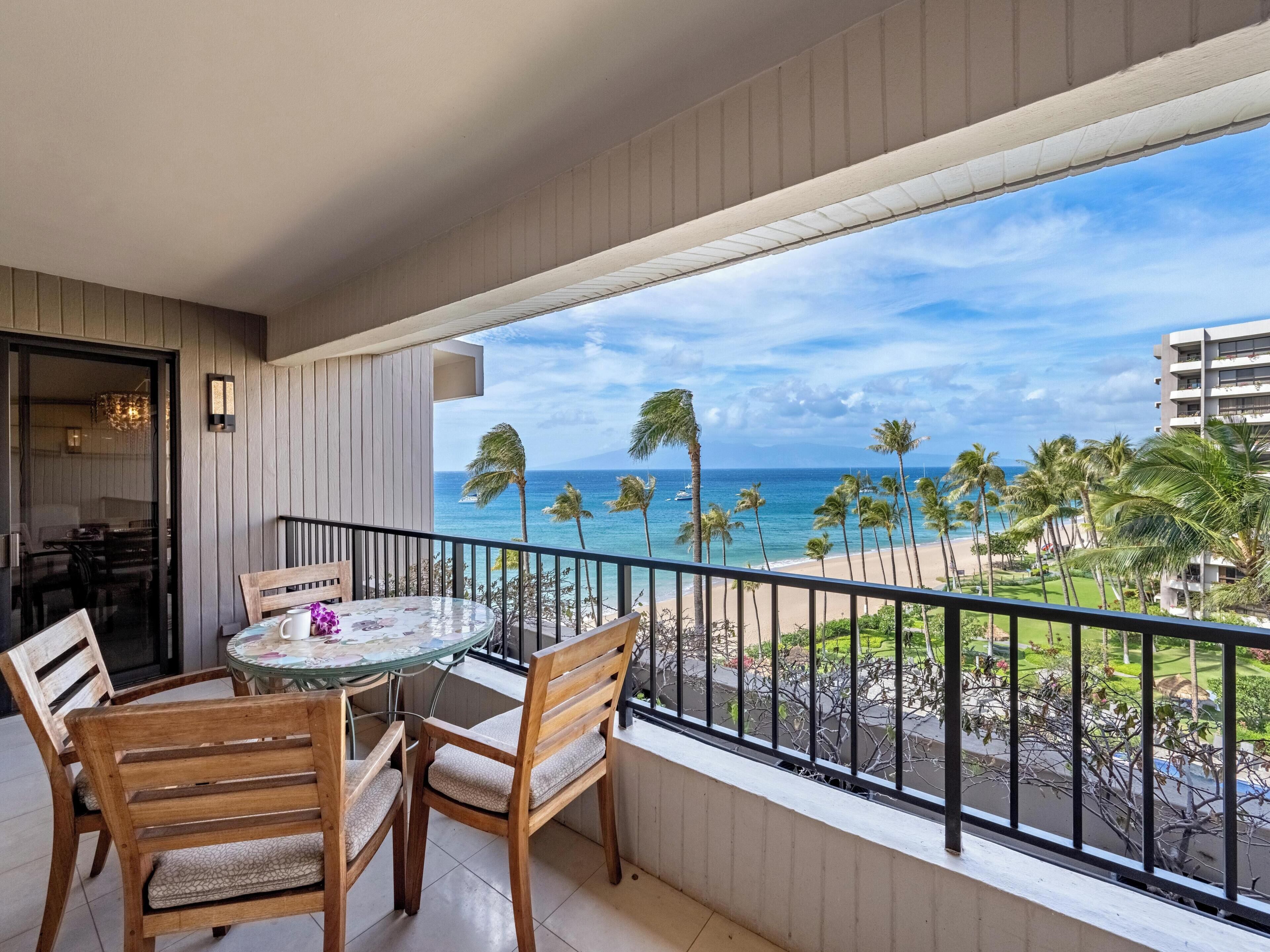 A balcony with a glass table and chairs, overlooking a beach with palm trees and a clear sky.