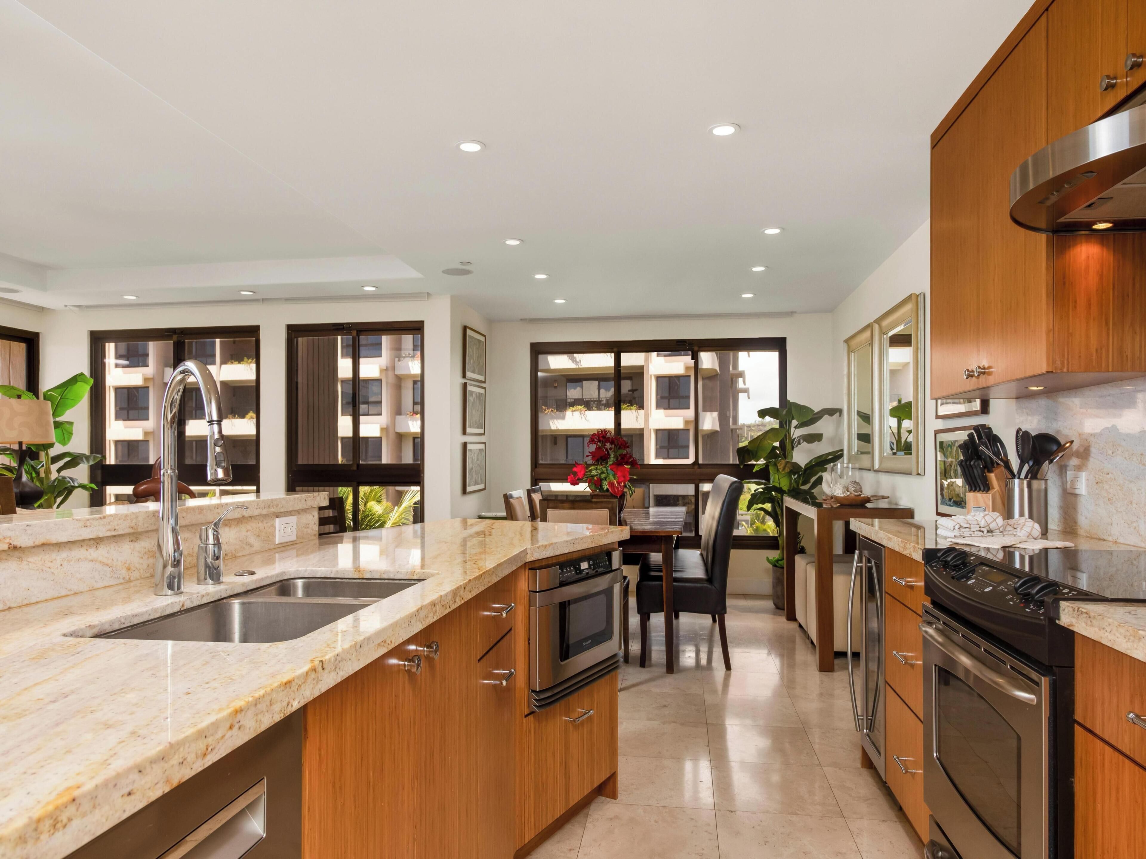 A modern kitchen with granite countertops, stainless steel appliances, wooden cabinets, and large windows offering views of a residential building.