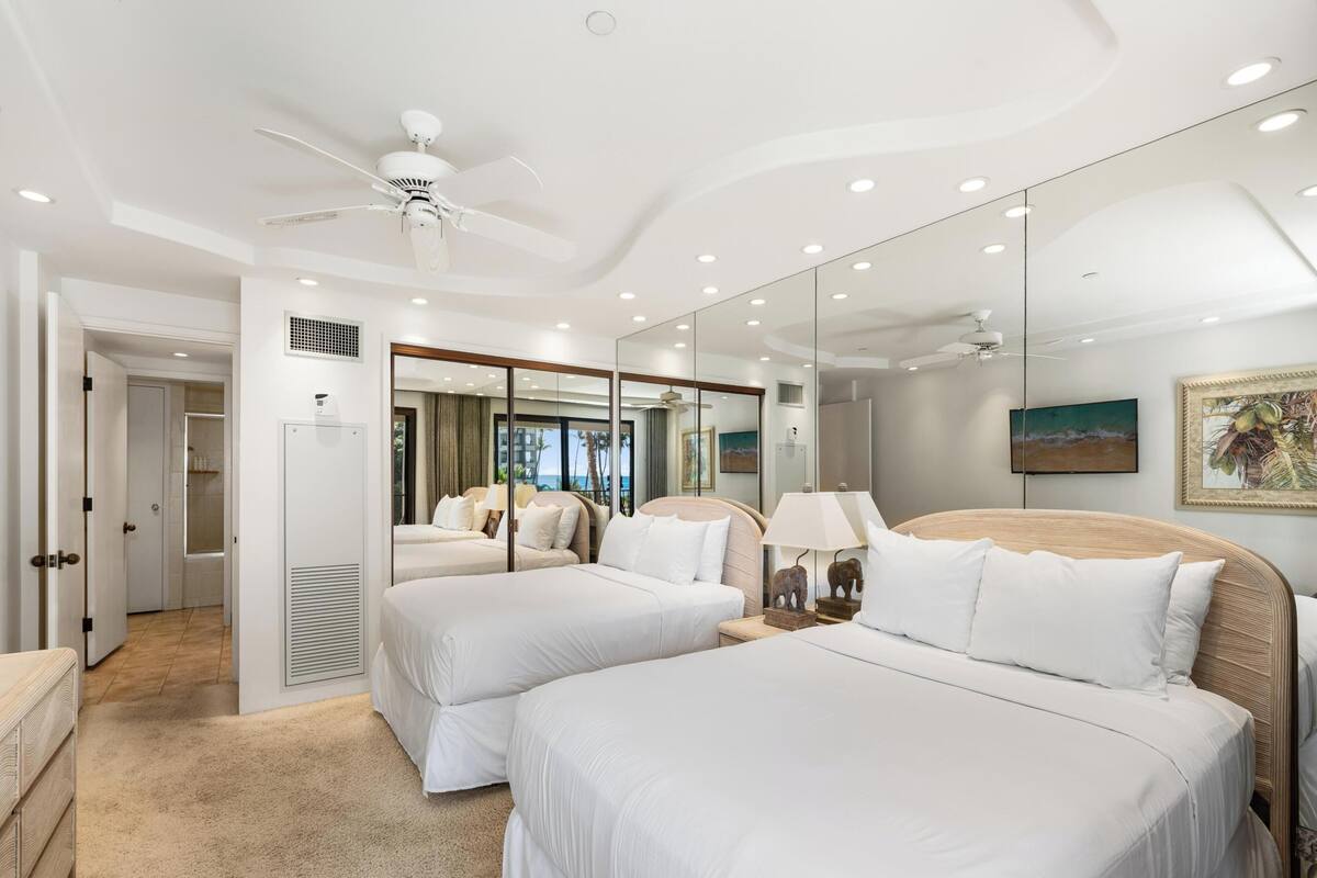 A hotel room with two beds, a ceiling fan, a large mirror, and a view of the ocean.