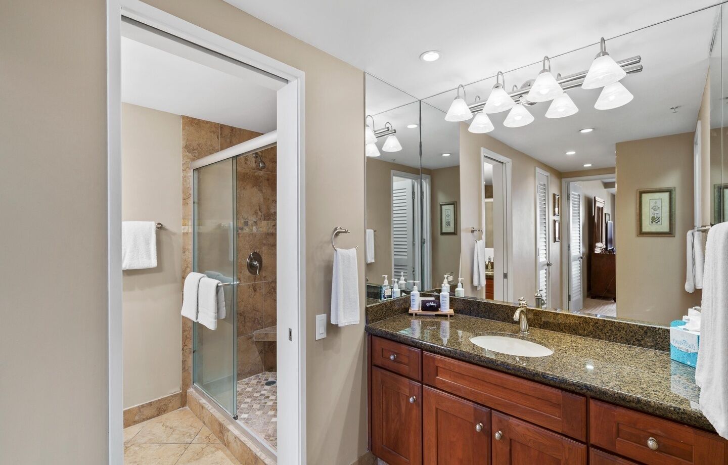 Une salle de bain moderne dotée d’une douche à parois de verre, d’un grand meuble-lavabo avec un plan de travail en granit et d’un miroir équipé de plusieurs lampes.