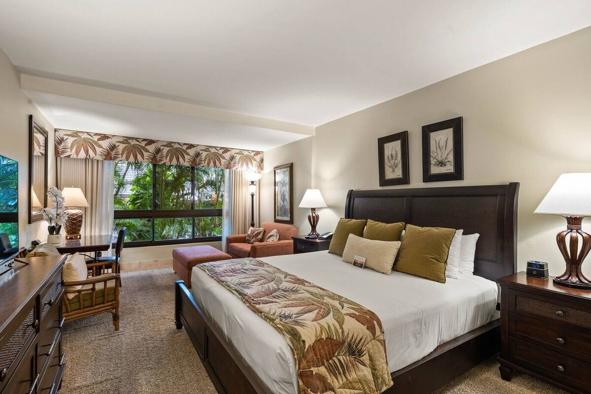 A hotel room with a large bed, a desk, a chair, and a window with a view of greenery.