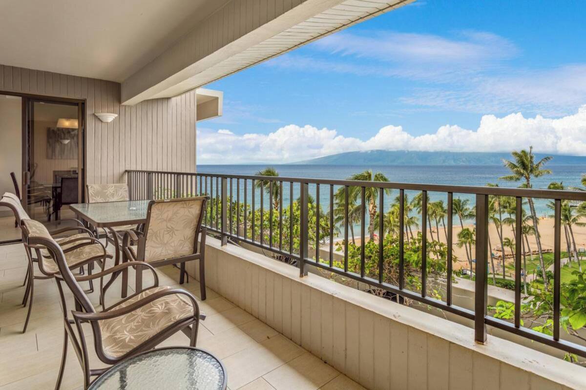A balcony with a table and chairs, overlooking a beach with palm trees and the ocean.