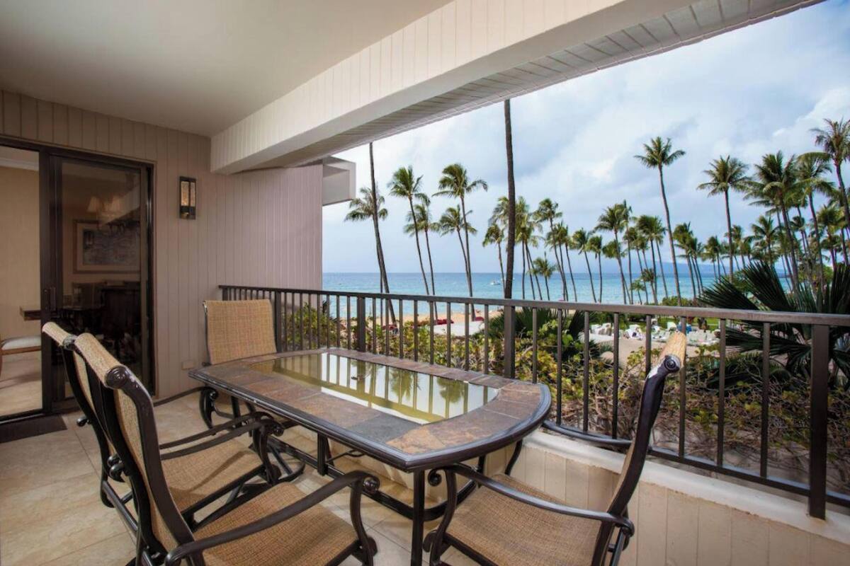 A balcony with a glass-top table and chairs, overlooking a beach with palm trees and the ocean.