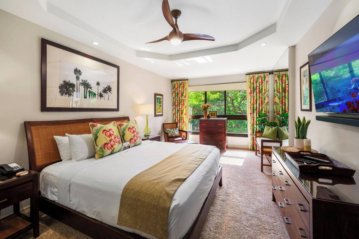 A hotel room with a large bed, a ceiling fan, a TV, a dresser, and a window with curtains.