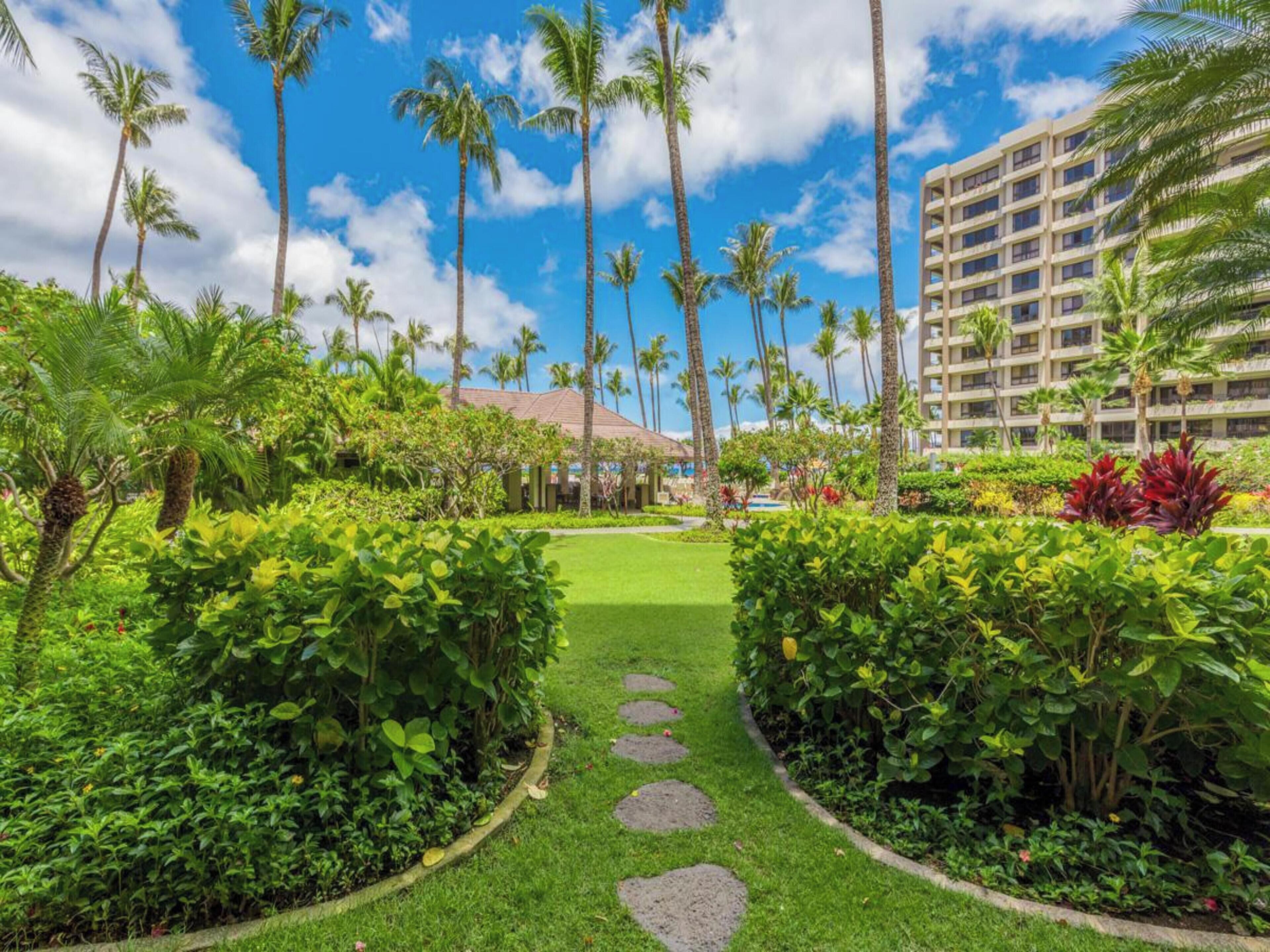 A lush garden pathway leading to a building with a red roof, surrounded by tall palm trees and dense greenery.